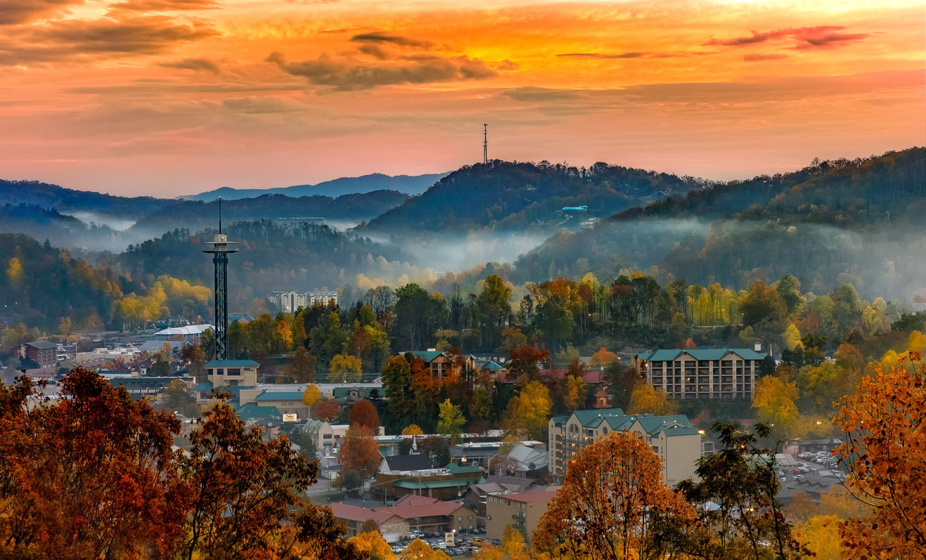 Autumn sunrise over one of the best fall towns in America, with colorful trees, charming buildings, a tower, mist drifting through the streets, and rolling hills in the background under an orange sky.