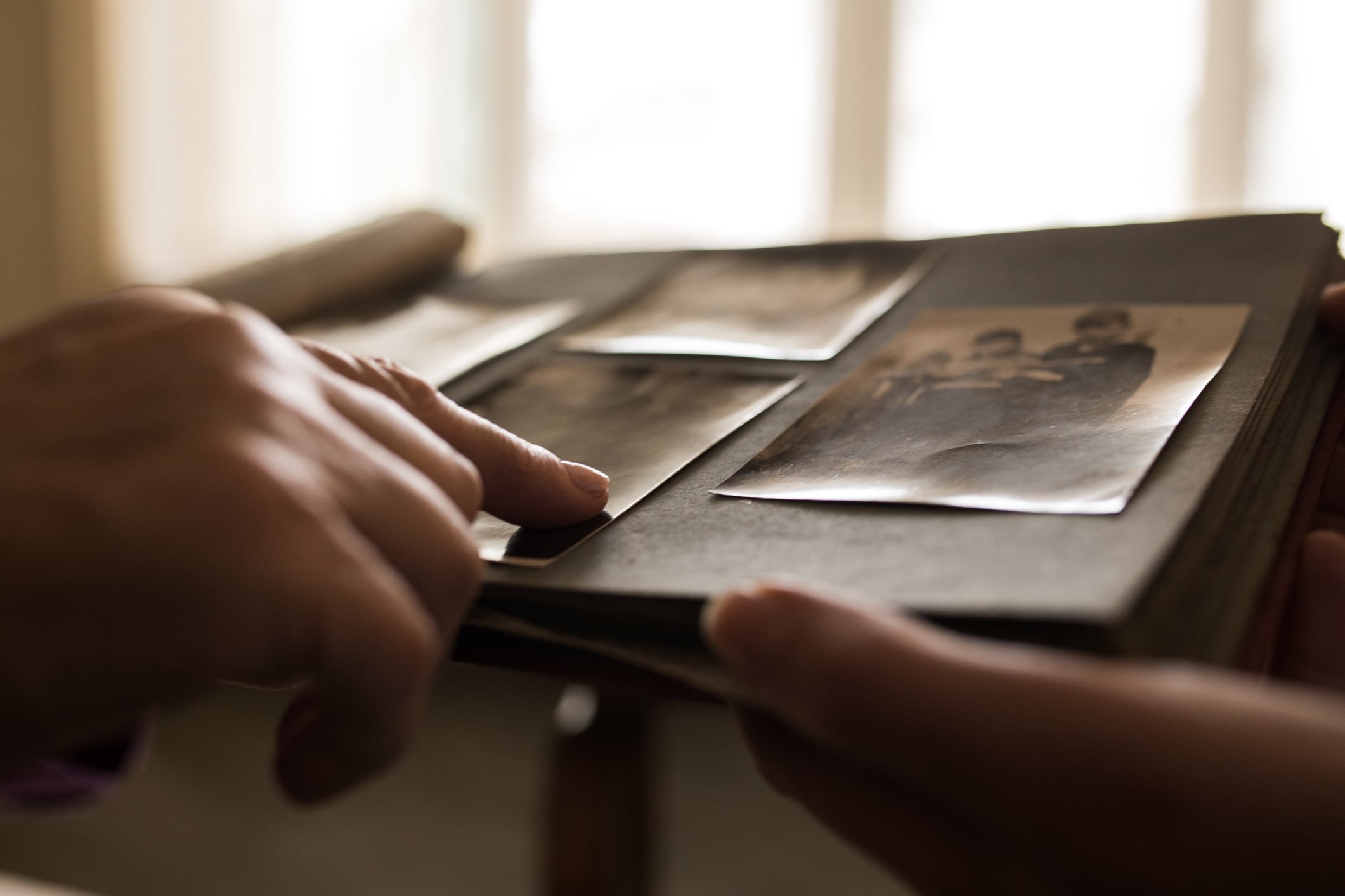 Two hands hold and point at old black-and-white photographs in a vintage photo album, with soft light coming from a window in the background, evoking cherished memories from days gone by, as if reminiscing about life in the 80s.