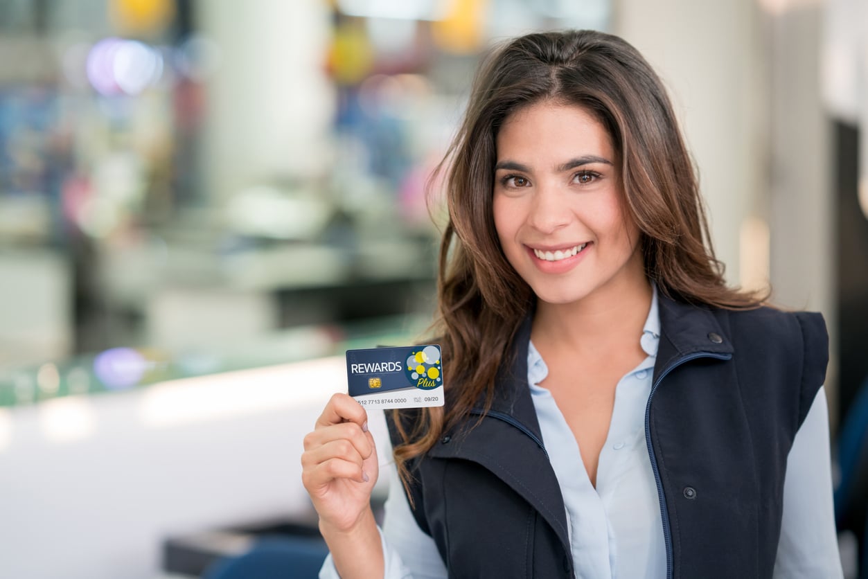 A smiling woman with long brown hair holds up a rewards card in a brightly lit indoor setting, wearing a blue shirt and dark vest, ready to put her travel habits to good use.