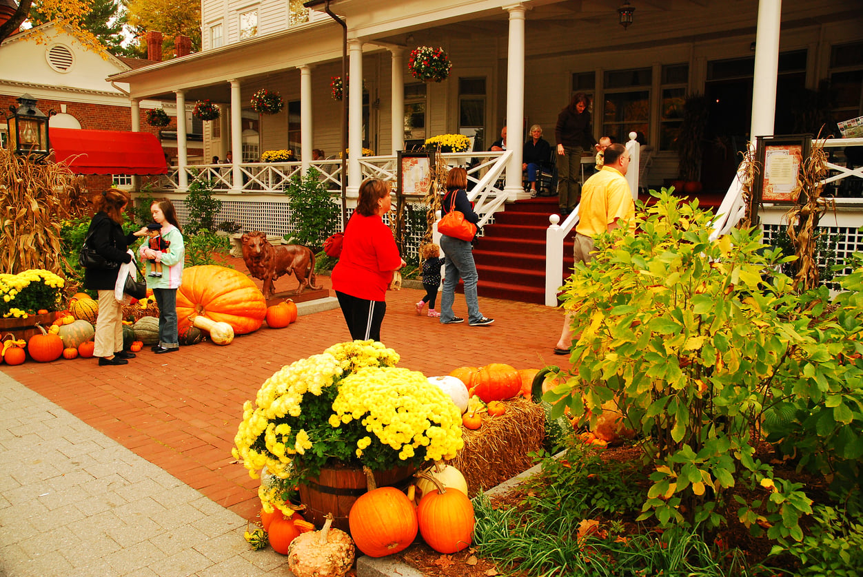 People stand outside a building decorated for fall with pumpkins, gourds, hay bales, and yellow flowers. Some visitors talk, and others walk up the red steps to the porch of one of the area’s charming historic hotels. Lush greenery and festive decor fill the scene.