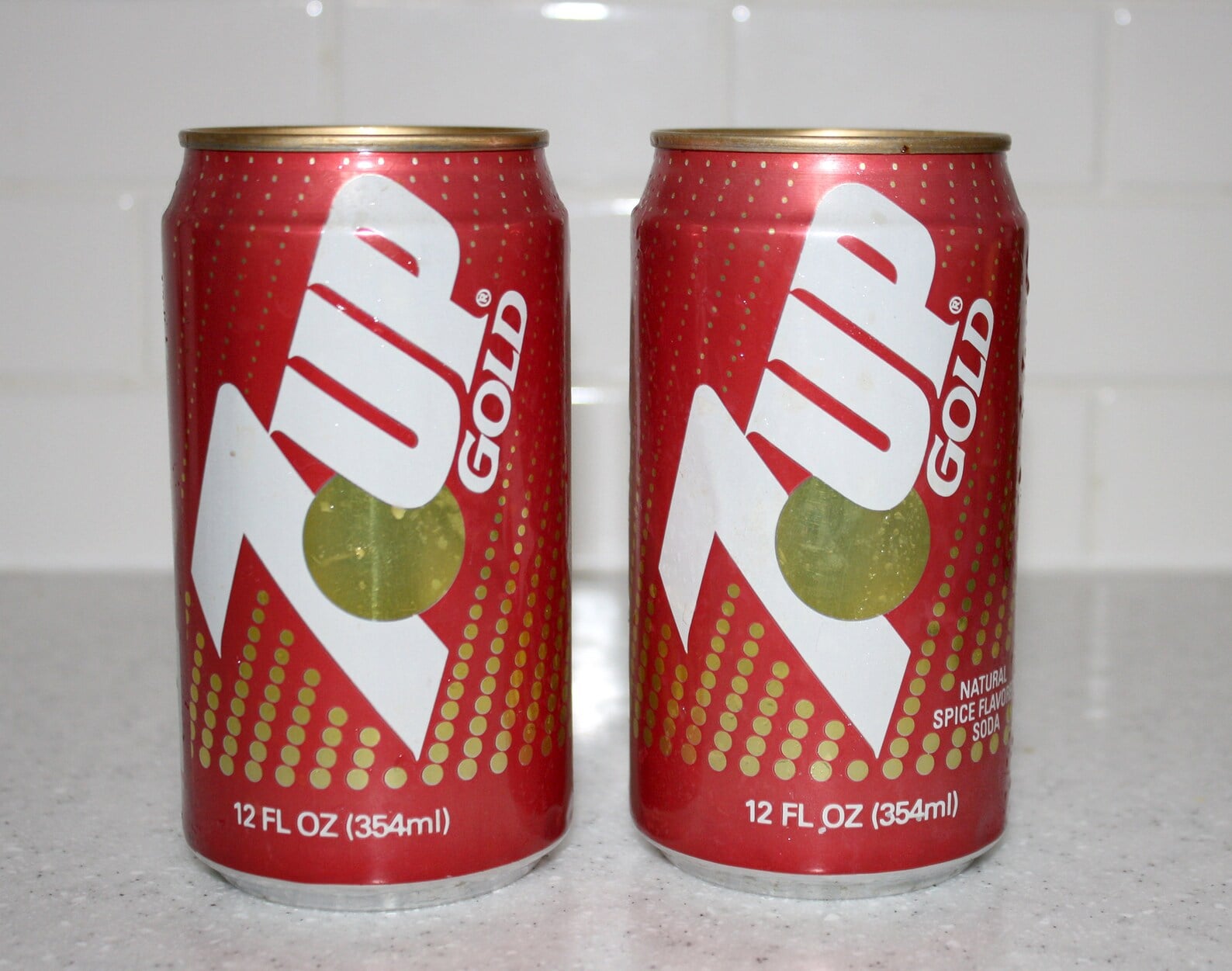Two red cans of 7UP Gold, each 12 fl oz (354 ml), are placed side by side on a light countertop with a white tiled background.