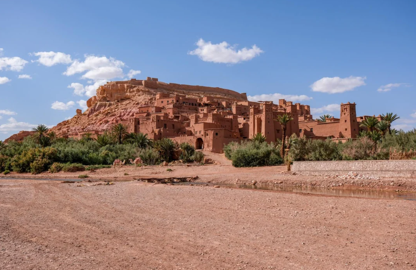 An ancient mud-brick fortress and village sit at the base of a rocky hill under a blue sky with scattered clouds, surrounded by green vegetation and a dry, sandy foreground.