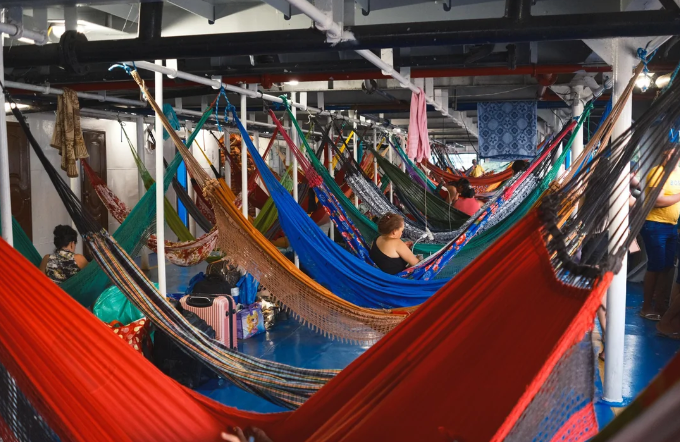 People relax in colorful hammocks hanging close together inside a crowded room, with bags and personal belongings nearby, under fluorescent lighting and exposed pipes.