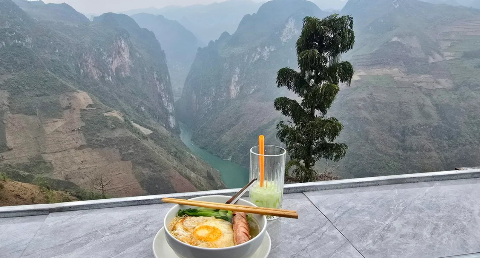 A bowl of noodles with an egg, sausage, and greens, plus a glass of green drink sit on a table overlooking a scenic mountain valley with a winding river below.