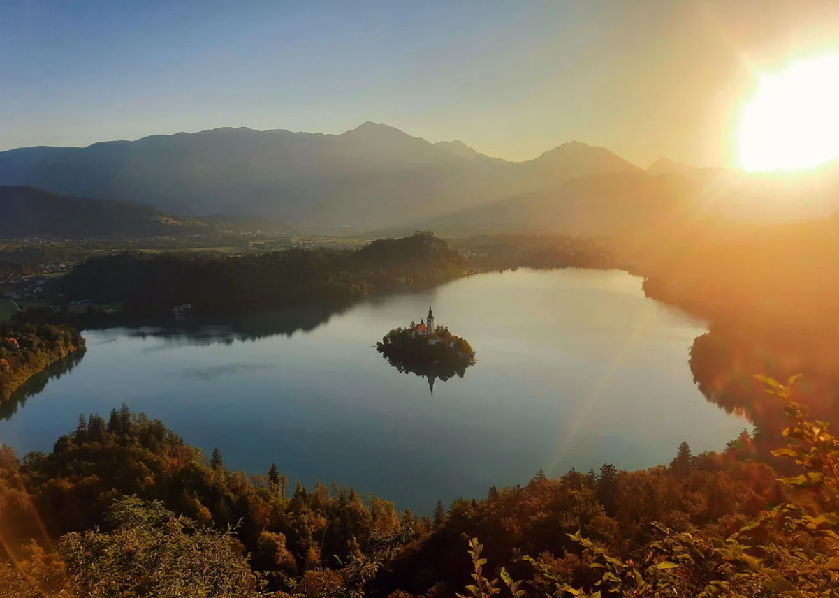 A serene lake with a small island and church in the center, surrounded by forested hills and mountains, with the sun setting in the background and warm light reflecting on the water.