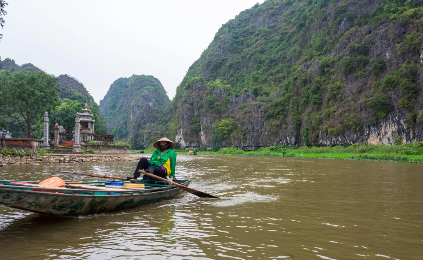A person in a traditional conical hat rows a wooden boat on a river surrounded by tall, green limestone mountains and lush vegetation.