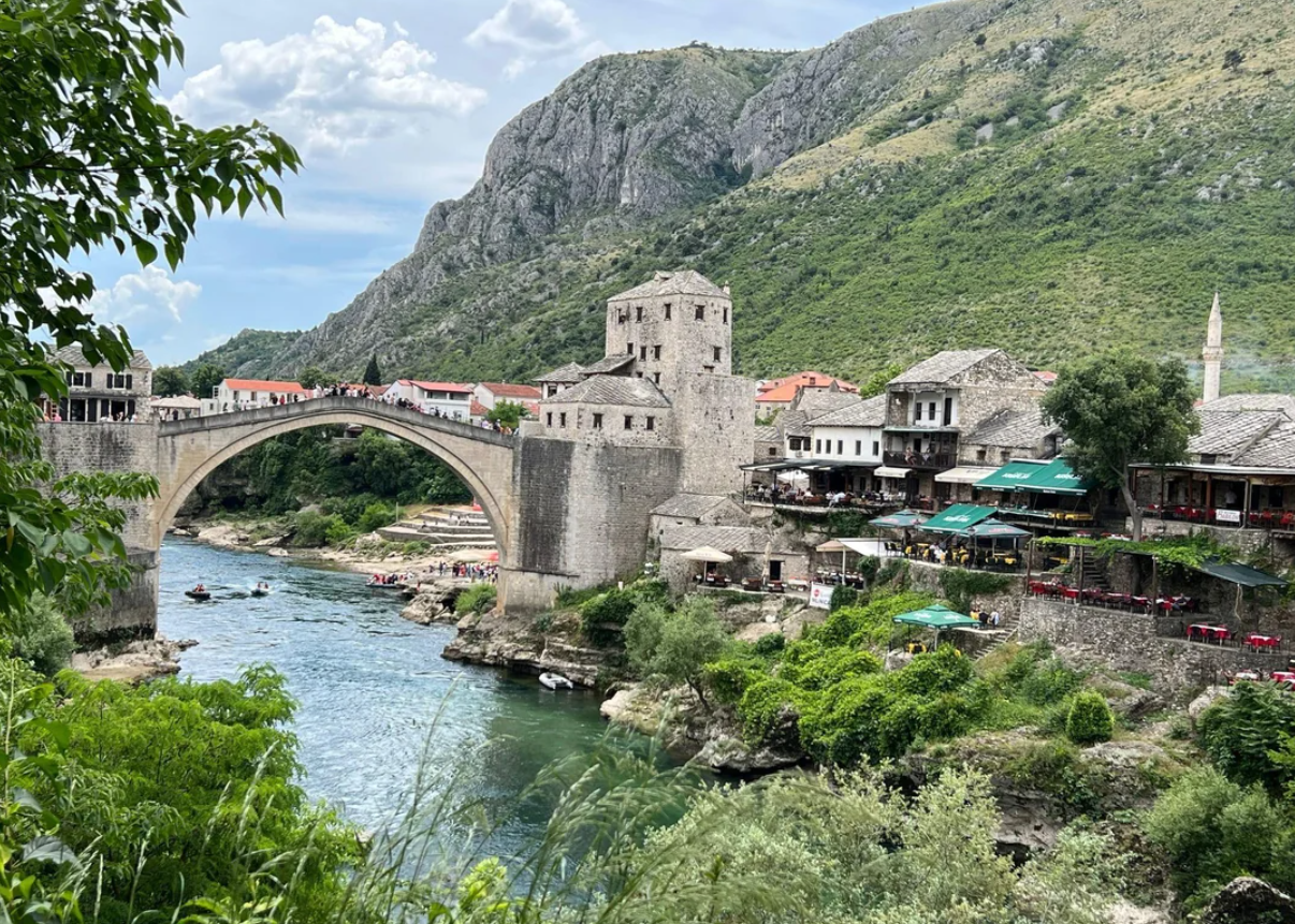 A stone arch bridge spans a river, surrounded by historic buildings and green hills under a partly cloudy sky. People walk across the bridge and relax at riverside cafes.