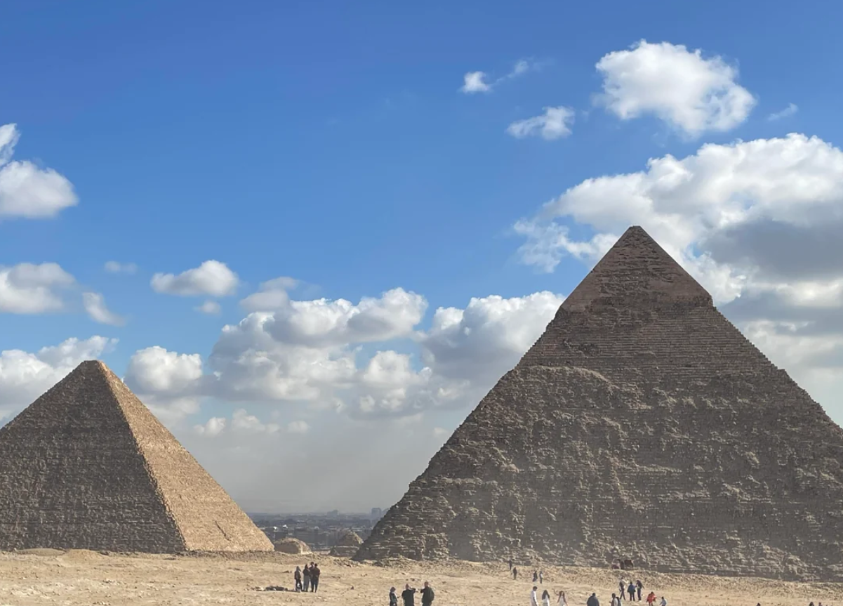 Two large pyramids stand under a blue sky with scattered clouds. People are gathered at the base, emphasizing the massive scale of the ancient structures. The landscape is dry and sandy.