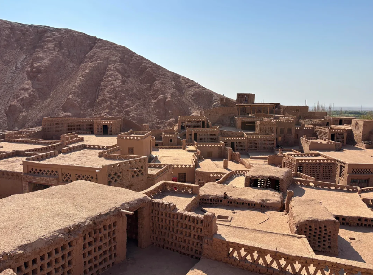 A cluster of mud-brick buildings with lattice walls sits at the base of a large, rocky hill under a clear blue sky. Desert landscape surrounds the ancient-looking settlement.