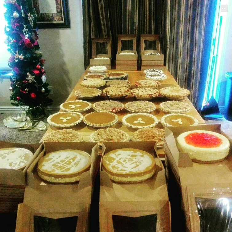 A long table filled with various pies and cheesecakes, with some boxed in the foreground. A decorated Christmas tree stands to the left, and three wooden chairs are visible at the end of the table.