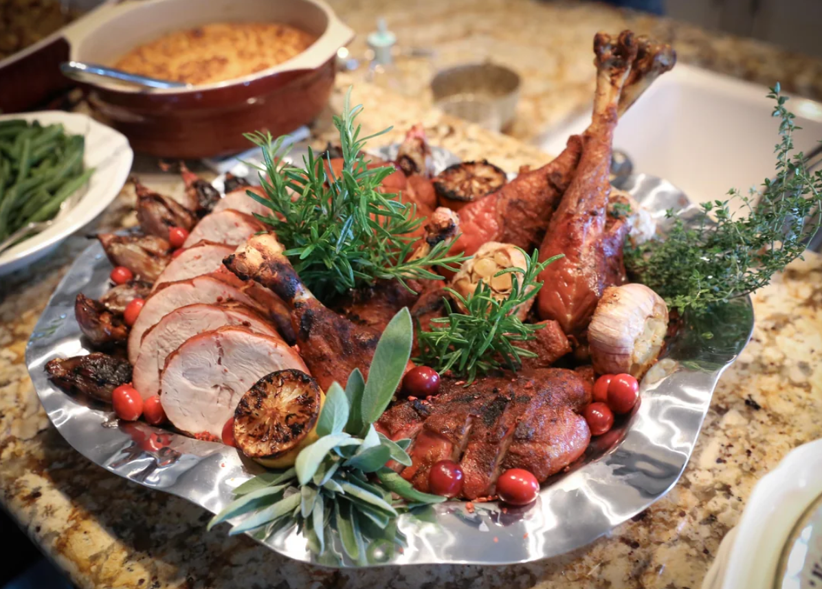 Sliced roasted turkey with drumsticks, herbs, cranberries, and grilled lemon on a platter, with green beans and casserole dishes in the background on a granite countertop.
