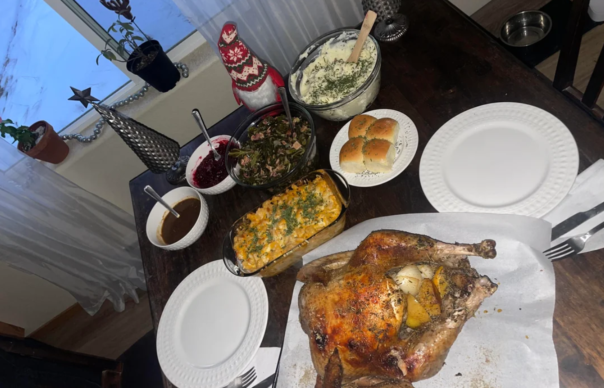 A roasted chicken with lemon slices sits on a table set with plates, dinner rolls, salad, a casserole, cranberry sauce, gravy, and a bowl of mashed potatoes, near a window with holiday decorations.
