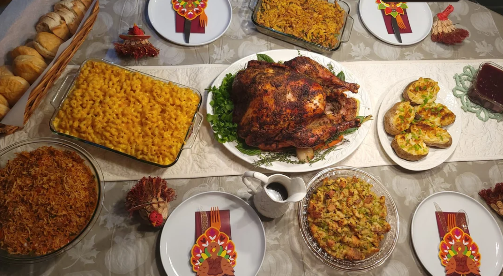 A Thanksgiving dinner table with roast turkey, macaroni and cheese, rice, stuffing, rolls, and cranberry sauce. Plates are set with decorative napkins and turkey-shaped napkin rings.