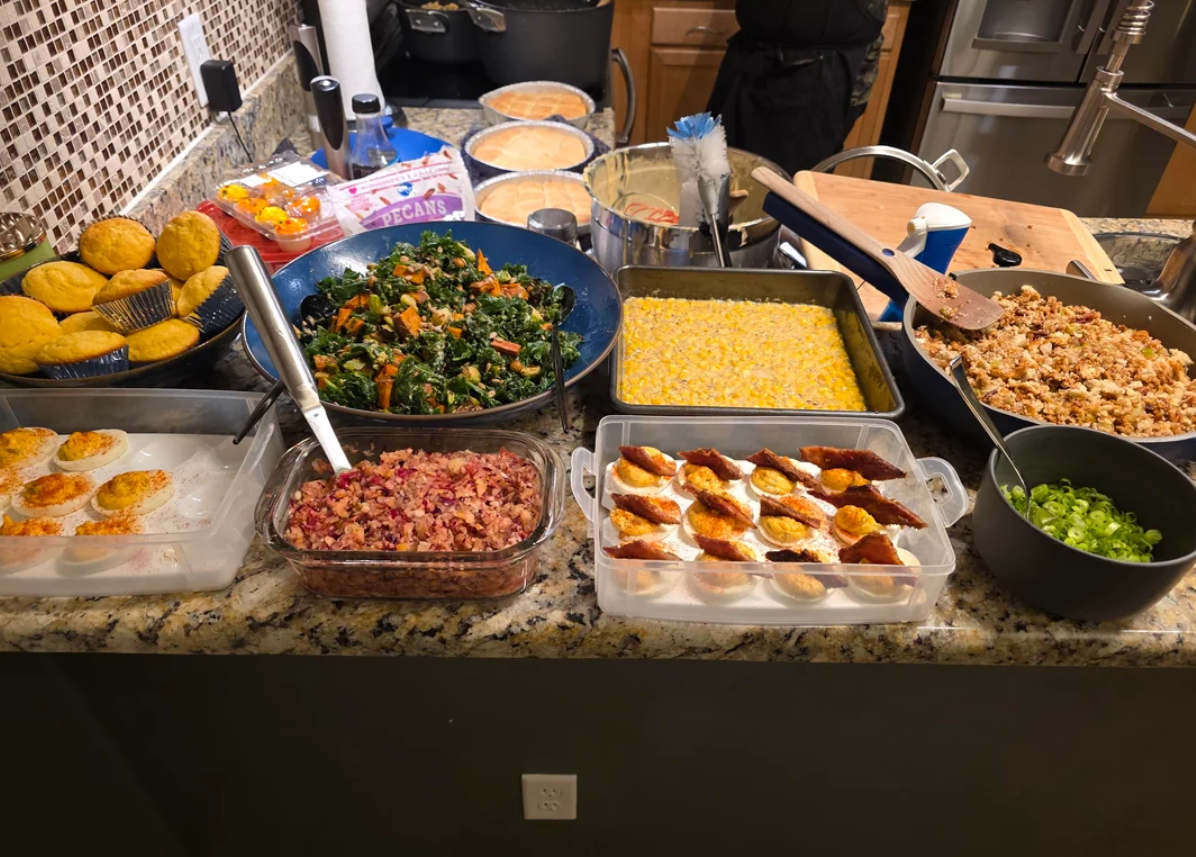 A kitchen counter filled with a variety of homemade dishes, including muffins, deviled eggs, greens, corn, dressing, salad, and desserts, ready for a meal.