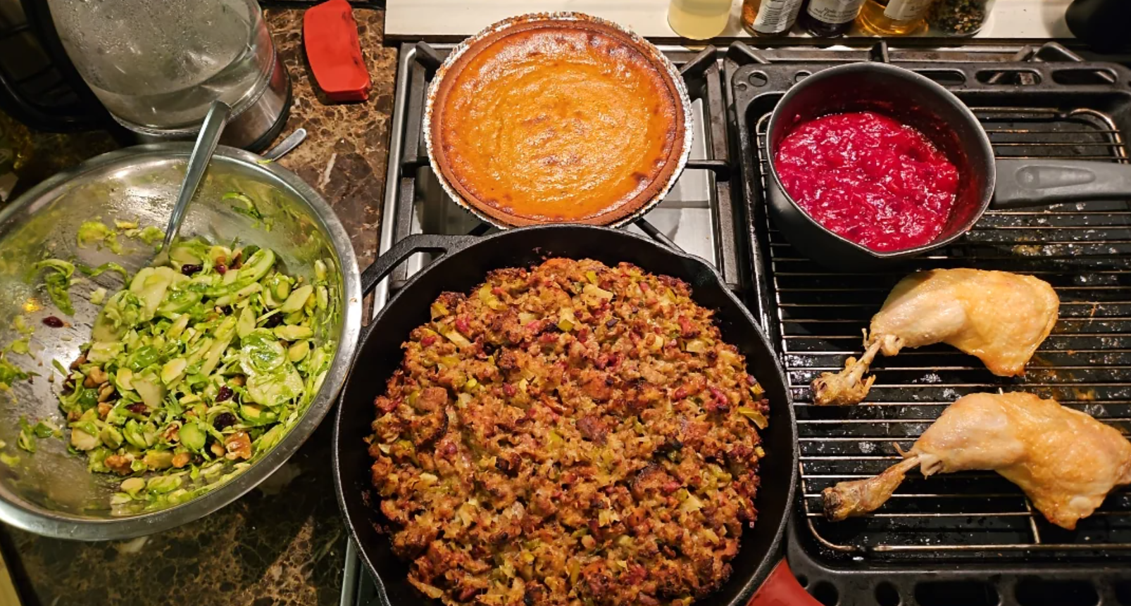 A bowl of Brussels sprouts, a skillet of stuffing, a sweet potato pie, a pot of cranberry sauce, and two baked chicken legs on a stovetop, all arranged for a holiday meal.