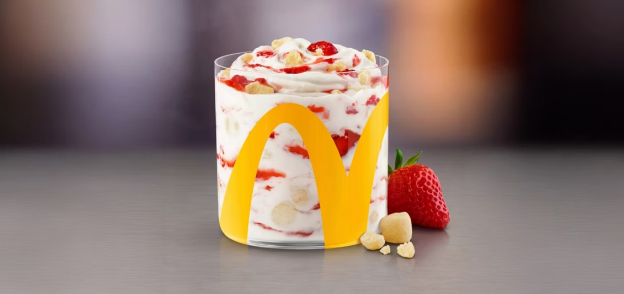 A McDonald’s dessert in a clear cup with the golden arches logo, featuring swirls of vanilla soft serve, strawberry pieces, and crumbled cookies. A whole strawberry and cookie pieces are beside the cup—one of those Wild Fast-Food Facts you might not know is how these creative treats continue to surprise fans worldwide.