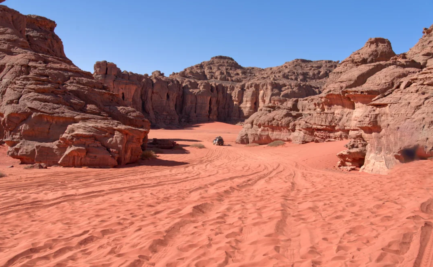 A vehicle drives across red sand dunes surrounded by tall, rugged rock formations under a clear blue sky.