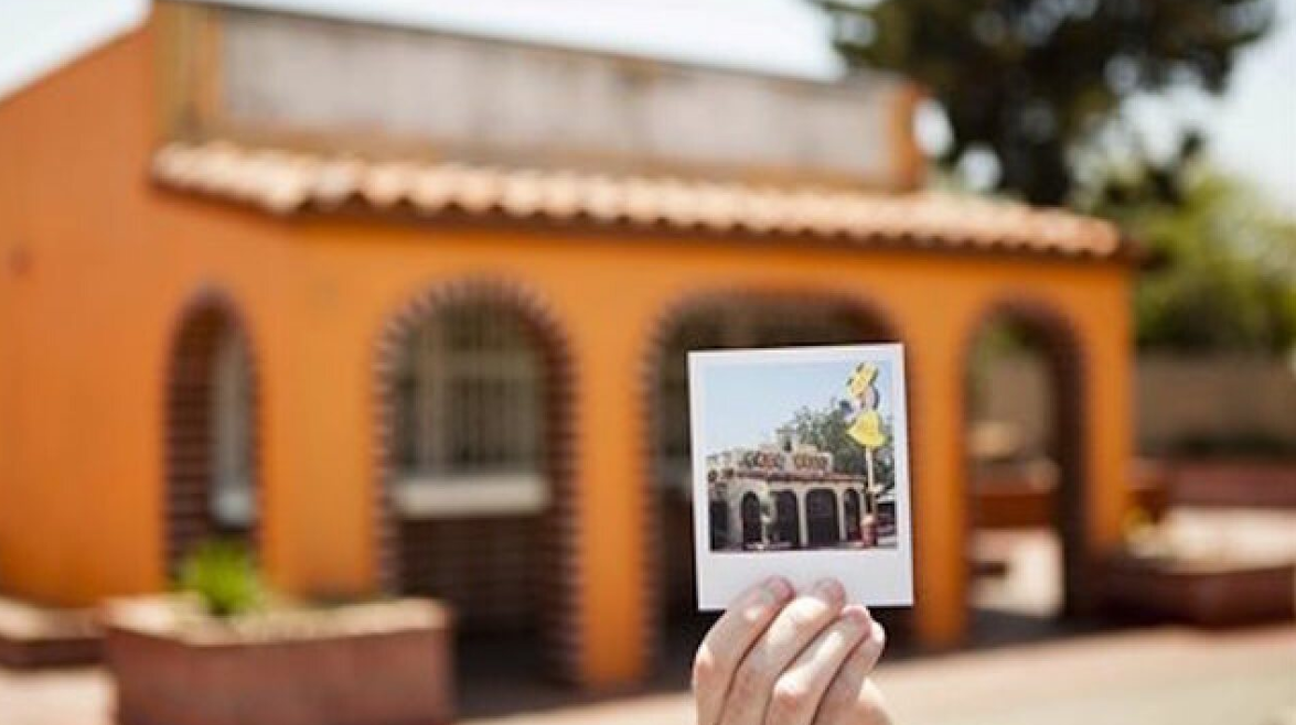 A hand holds a photo of a building in front of the same orange building, aligning the photo with the real structure in the background—much like discovering Wild Fast-Food Facts that perfectly match their surprising reality.
