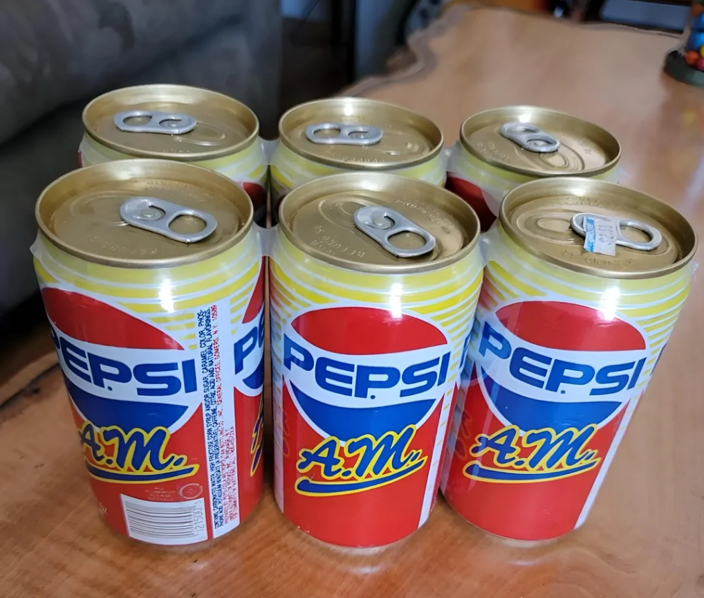 Six Soda Pops—Pepsi A.M. cans with yellow accents—are arranged on a wooden table, opened and grouped with their pull tabs facing up. A couch is visible in the background.