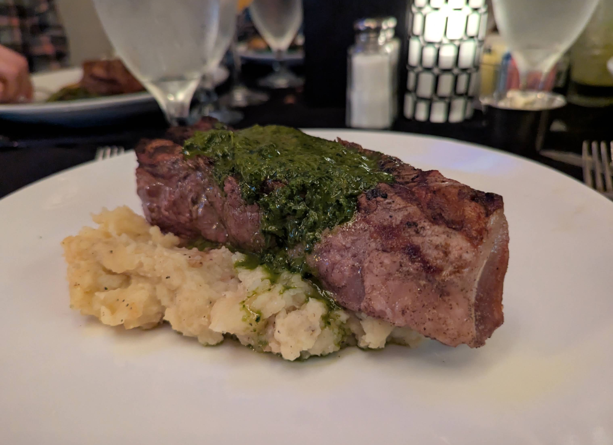 A grilled steak topped with green chimichurri sauce sits on a mound of mashed potatoes, served on a white plate with water glasses and a salt shaker in the background.