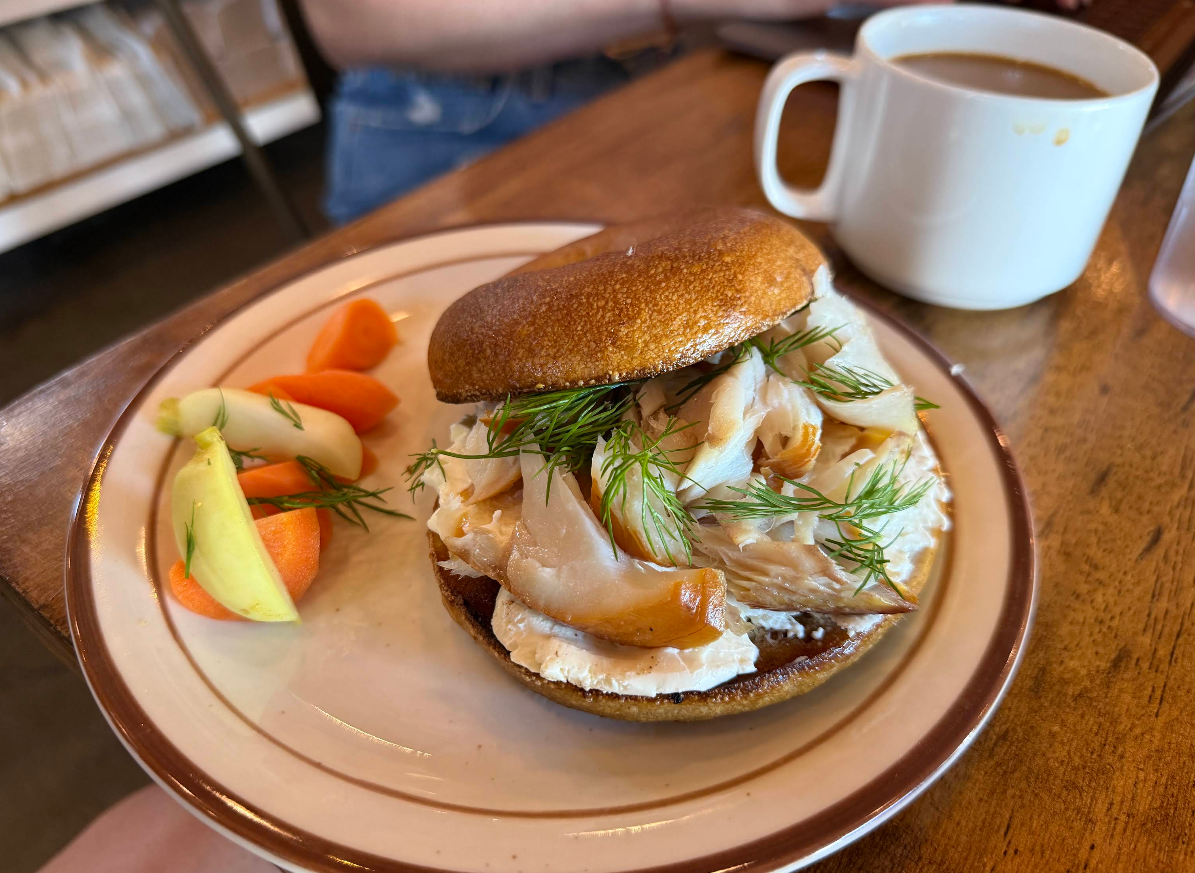 A bagel with cream cheese, smoked fish, and fresh dill is on a plate with carrot and cucumber slices. A white mug of coffee sits nearby on a wooden table.