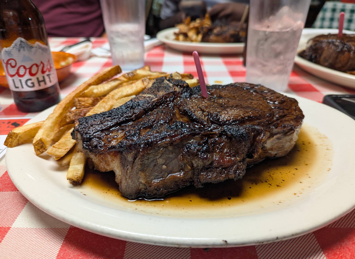 A plate with a grilled steak, French fries, and juice, on a red and white checkered table next to a Coors Light bottle and a water glass.