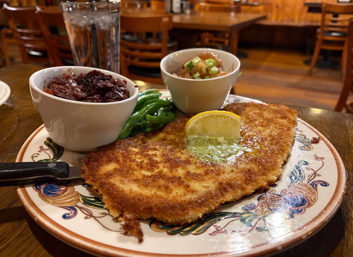 A plate with breaded schnitzel topped with a lemon wedge and green beans, served with bowls of red cabbage and potatoes, and a glass of water in the background on a wooden table.