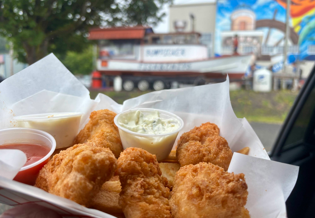 A basket of golden fried fish and fries with tartar sauce and ketchup, held up in front of a seafood restaurant and a boat in the background.