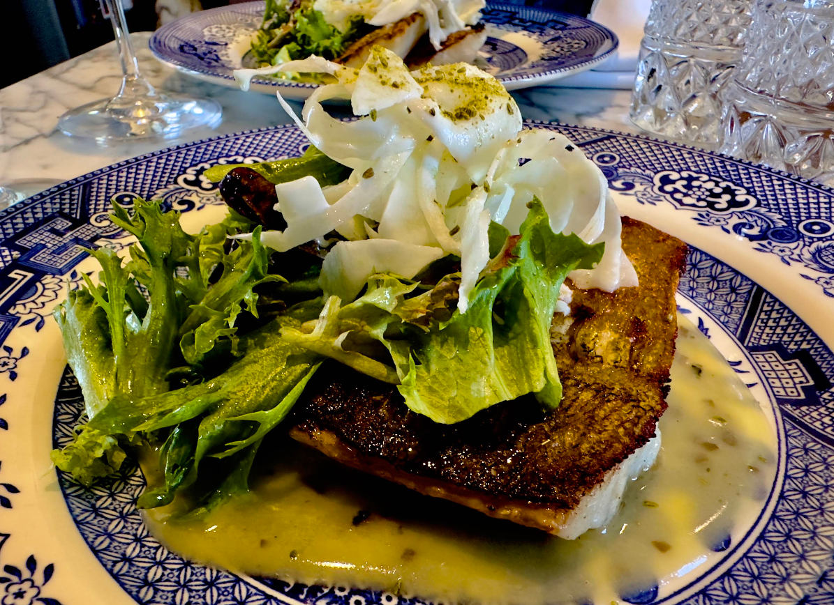 A plated dish featuring seared fish with crispy skin, topped with fresh mixed greens and thinly sliced fennel, served with a creamy sauce on a blue and white patterned plate.