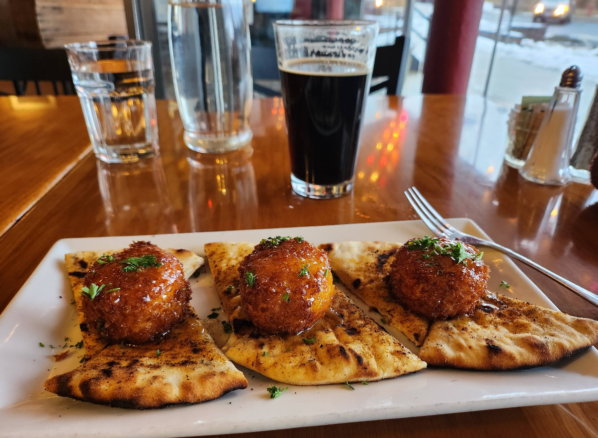 Three pieces of grilled flatbread topped with crispy, round appetizers and herbs are served on a rectangular white plate, with a glass of dark beer, water, and utensils on a wooden table in a restaurant.