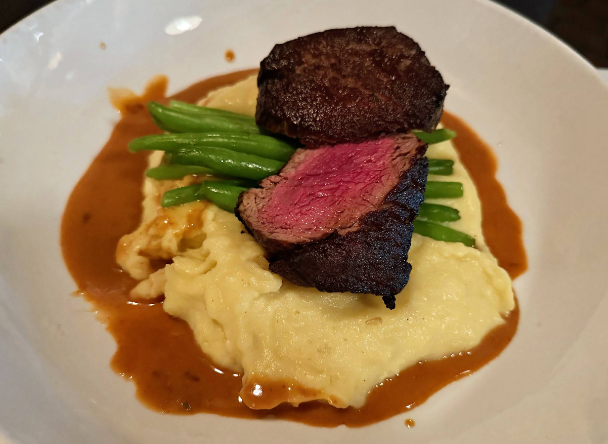 Sliced medium-rare steak atop mashed potatoes with green beans, surrounded by brown gravy, served on a white plate.