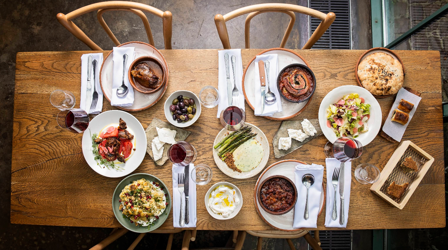 A wooden table set for four with wine, bread, cheese, olives, various salads, dips, baked dishes, and desserts arranged neatly for a shared meal.