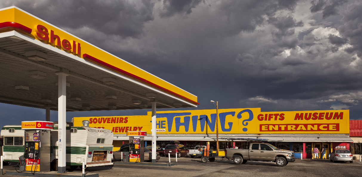 A Shell gas station with an attached souvenir and gift shop. A large yellow sign reads “THE THING? GIFTS MUSEUM ENTRANCE.” Cars and an RV are parked outside under a dramatic, cloudy sky.