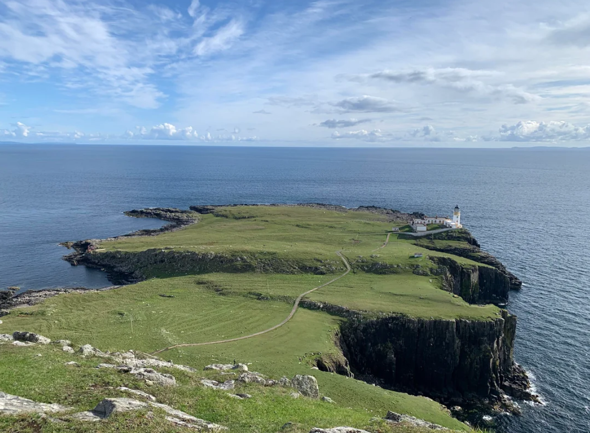 A grassy cliffside juts into the sea under a blue sky with scattered clouds; a small white lighthouse and buildings sit near the edge overlooking the ocean.