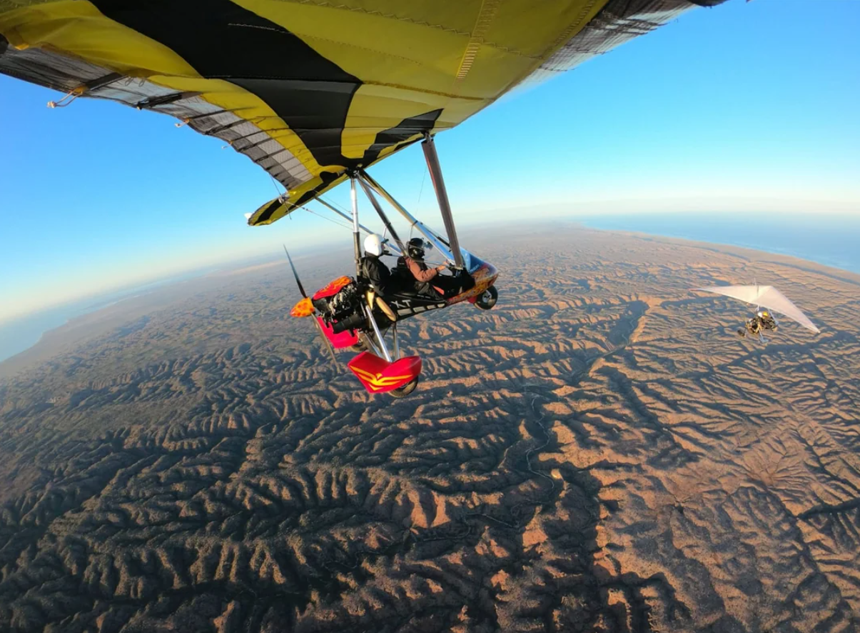 Two people fly a motorized hang glider high above a vast, rugged landscape. Another glider is visible in the distance under a clear blue sky.