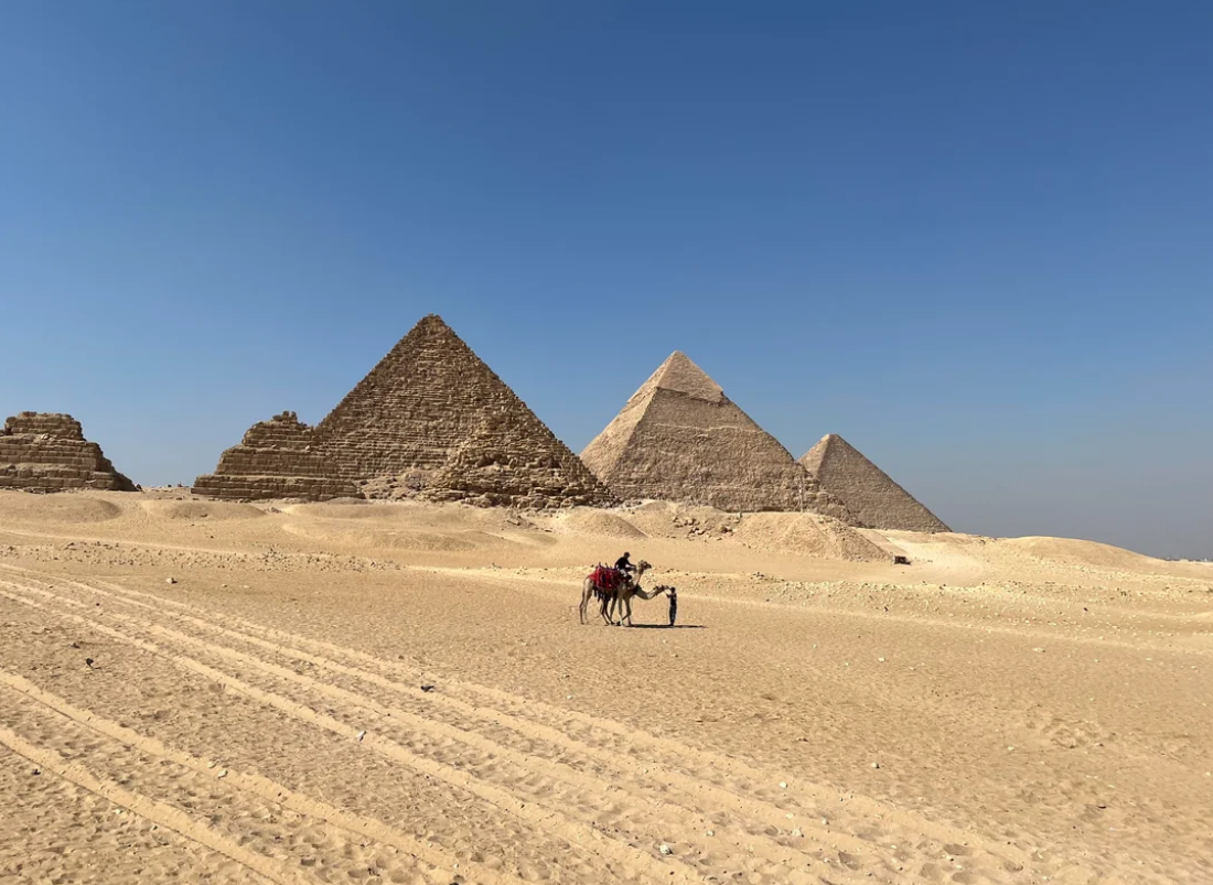 A person rides a camel in the sandy desert in front of the Pyramids of Giza, under a clear blue sky. Four pyramids are prominently visible in the background.