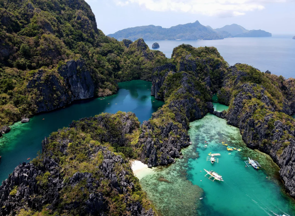 Aerial view of emerald and turquoise lagoons surrounded by rocky cliffs and lush greenery, with traditional boats floating on the water and distant mountains under a partly cloudy sky.