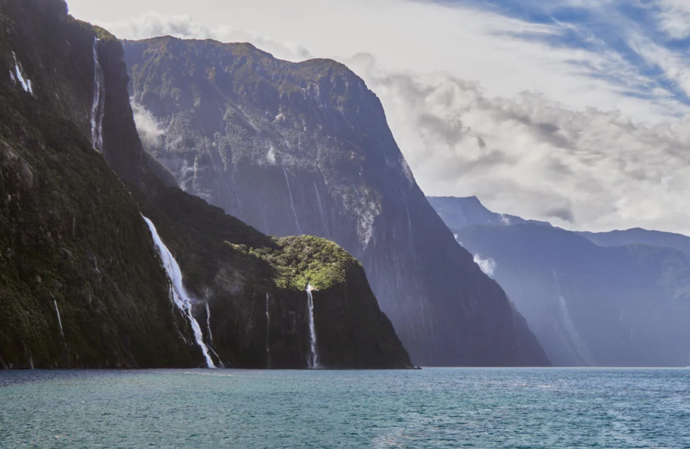 Tall, misty cliffs with several waterfalls cascade into a blue fjord under a partly cloudy sky, inviting awe and wonder for those who embark on travelers’ journeys, with distant mountains fading into the background.