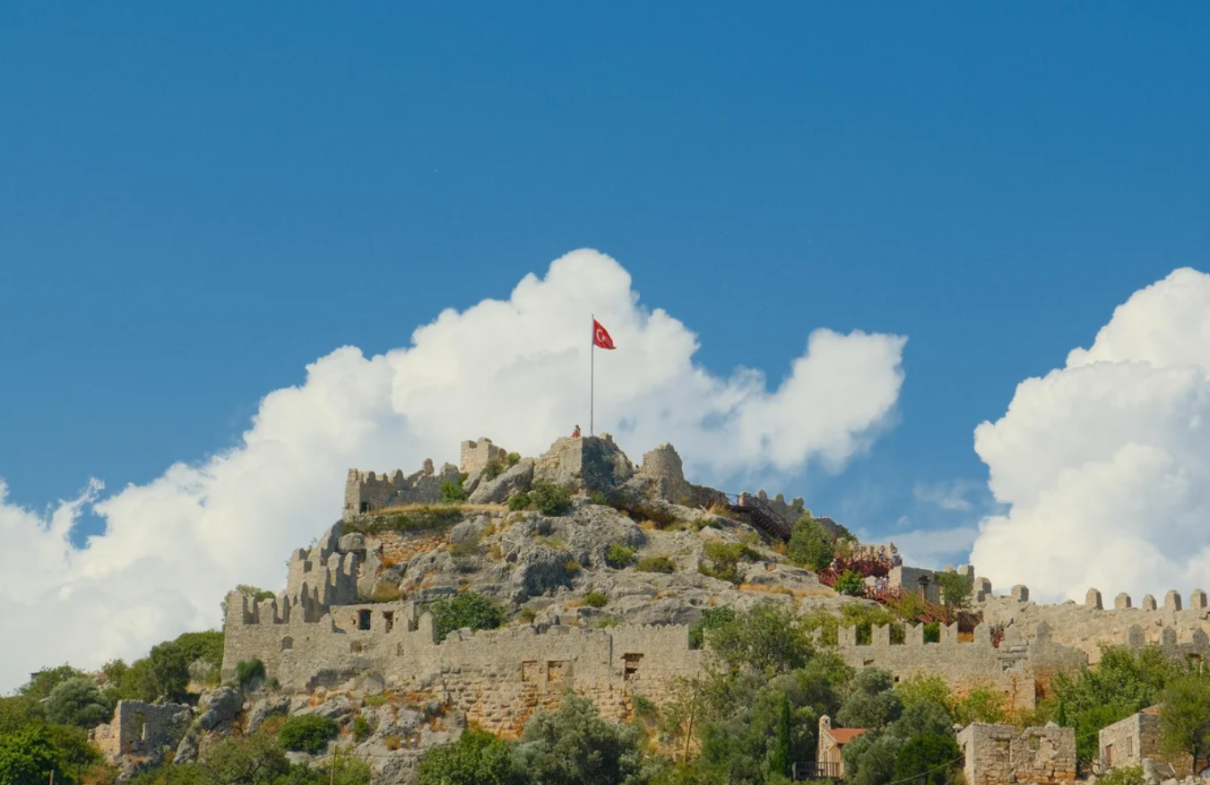 Ancient stone fortress ruins on a rocky hilltop with a Turkish flag flying at the summit, under a blue sky with scattered white clouds.