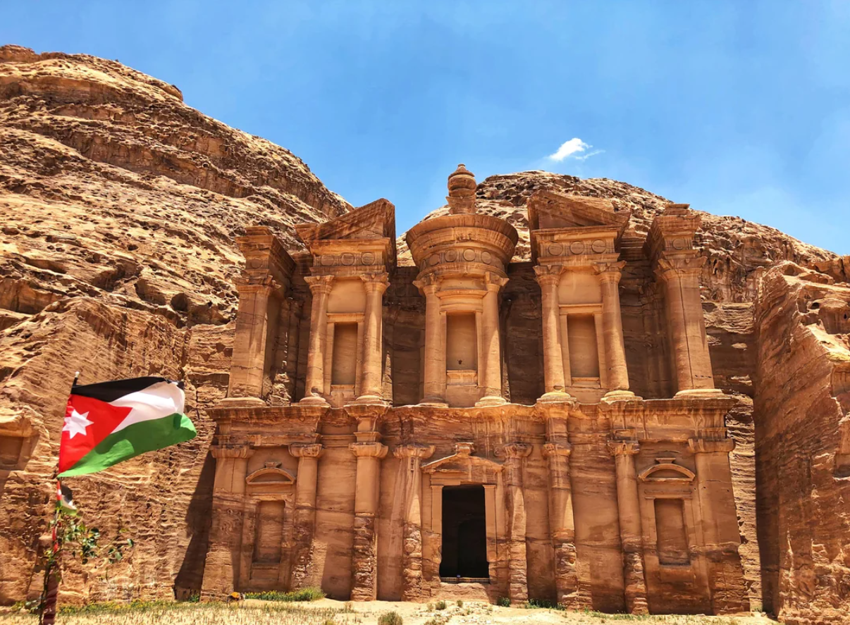 The Monastery, an ancient rock-cut structure in Petra, Jordan, stands under a blue sky. A Jordanian flag waves in the foreground as rocky cliffs surround the monument, inviting travelers’ journeys to explore its timeless beauty.
