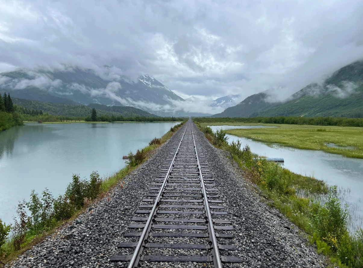 Railroad tracks stretch into the distance between two calm lakes, offering a tranquil route for Travelers’ Journeys, surrounded by green vegetation and mountains partially covered by clouds on a cloudy day.
