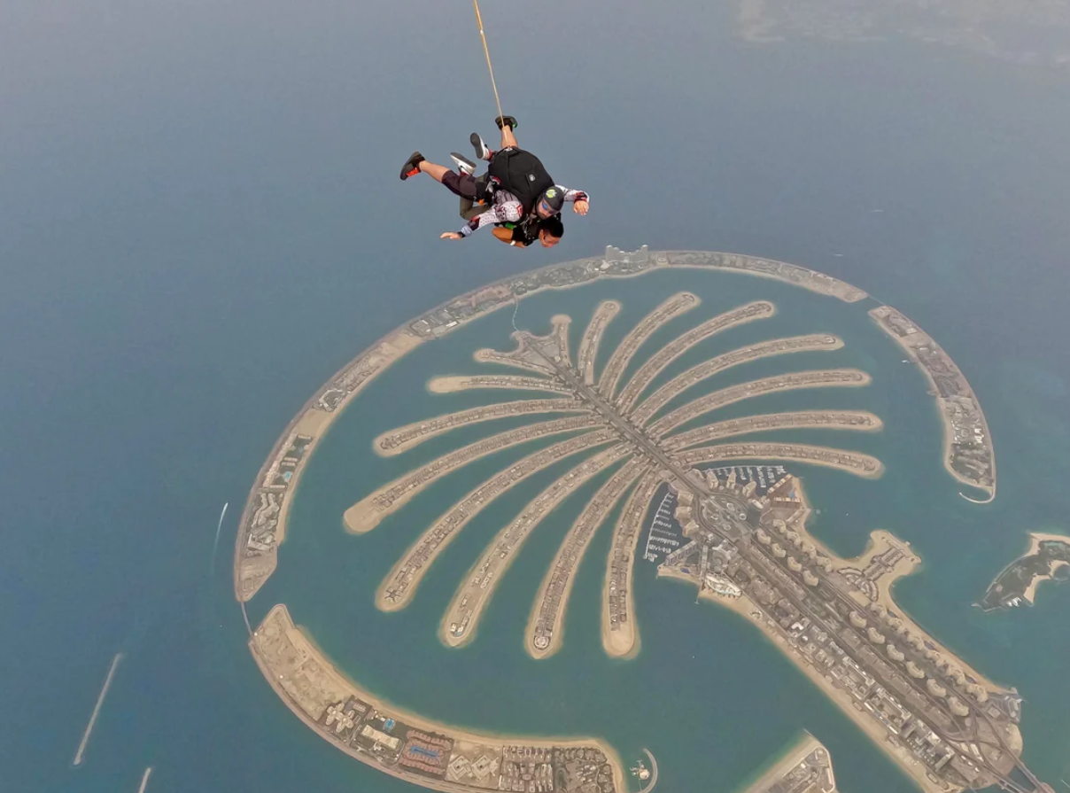 Two people tandem skydiving above Dubai’s Palm Jumeirah, an artificial island shaped like a palm tree, experience the thrill of the city from a unique perspective. Their descent encapsulates the spirit of Travelers’ Journeys, with blue water and distant city structures completing this unforgettable adventure.
