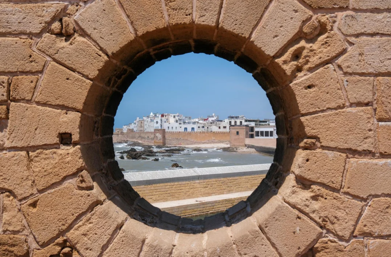 A round stone window frames a coastal city with white buildings, blue sky, and waves crashing on rocks along the shore—a picturesque scene inviting travelers’ journeys to explore its timeless beauty.