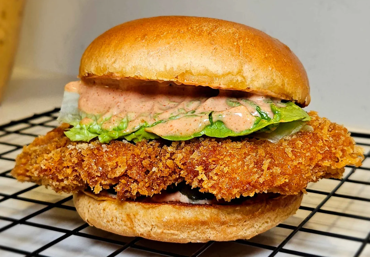 A crispy fried chicken sandwich with lettuce and creamy sauce in a toasted bun, placed on a cooling rack.