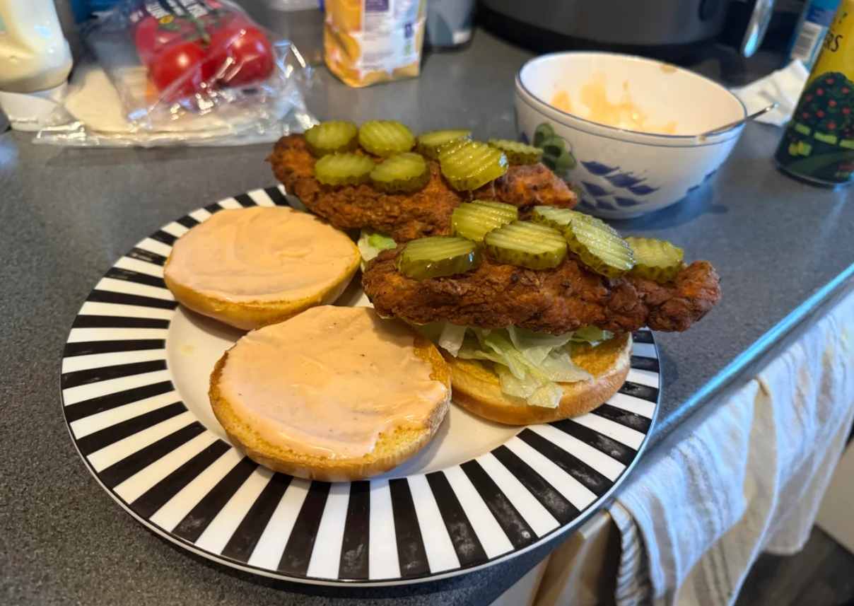 A plate holds two open fried chicken sandwiches with lettuce, pickle slices, and sauce on buns. Kitchen items, like tomatoes and a bowl, are visible in the background.