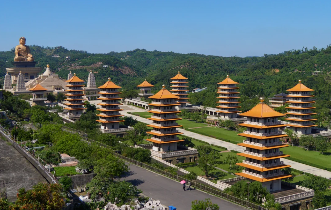 Aerial view of Fo Guang Shan Monastery in Taiwan with multiple orange-roofed pagodas, a large seated Buddha statue, lush greenery, and mountains in the background under a clear blue sky.