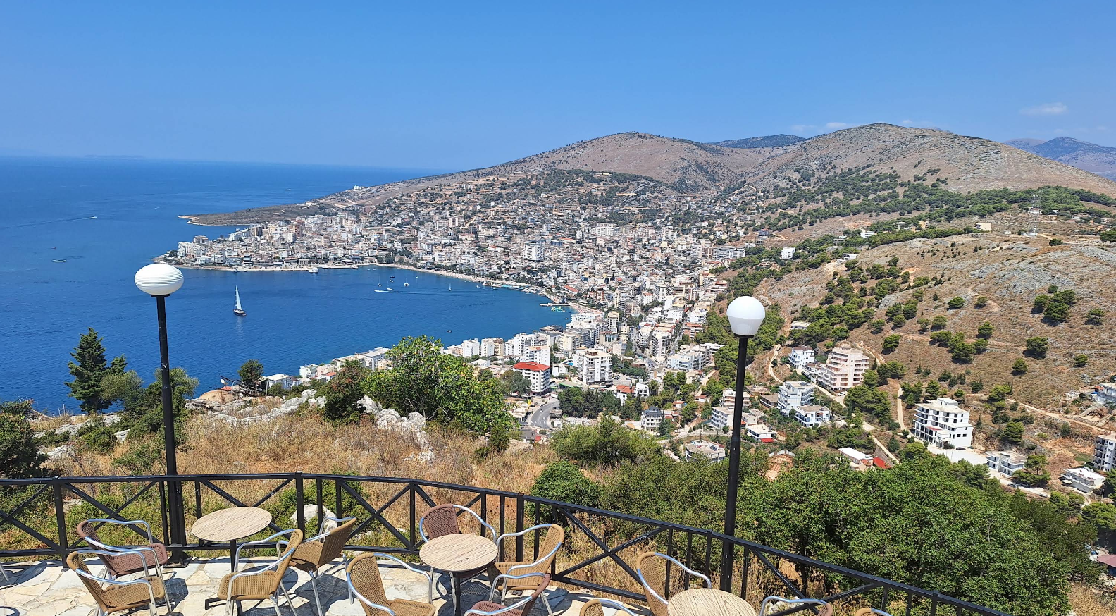 A seaside city with white buildings stretches along a bay, surrounded by hills. Outdoor café tables and chairs are in the foreground, with lampposts and greenery under a clear blue sky.