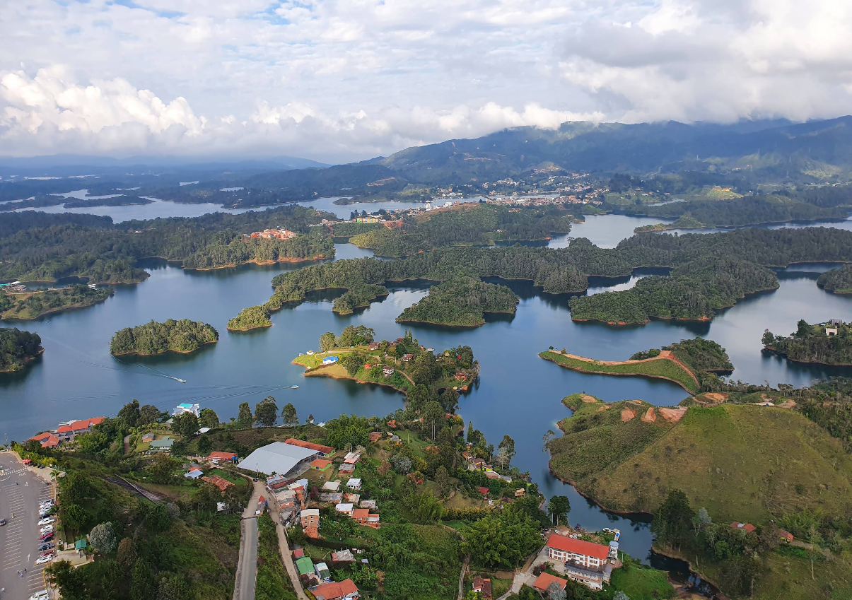 Aerial view of a lake with many small, lush islands, surrounded by hills, houses, and a partly cloudy sky.