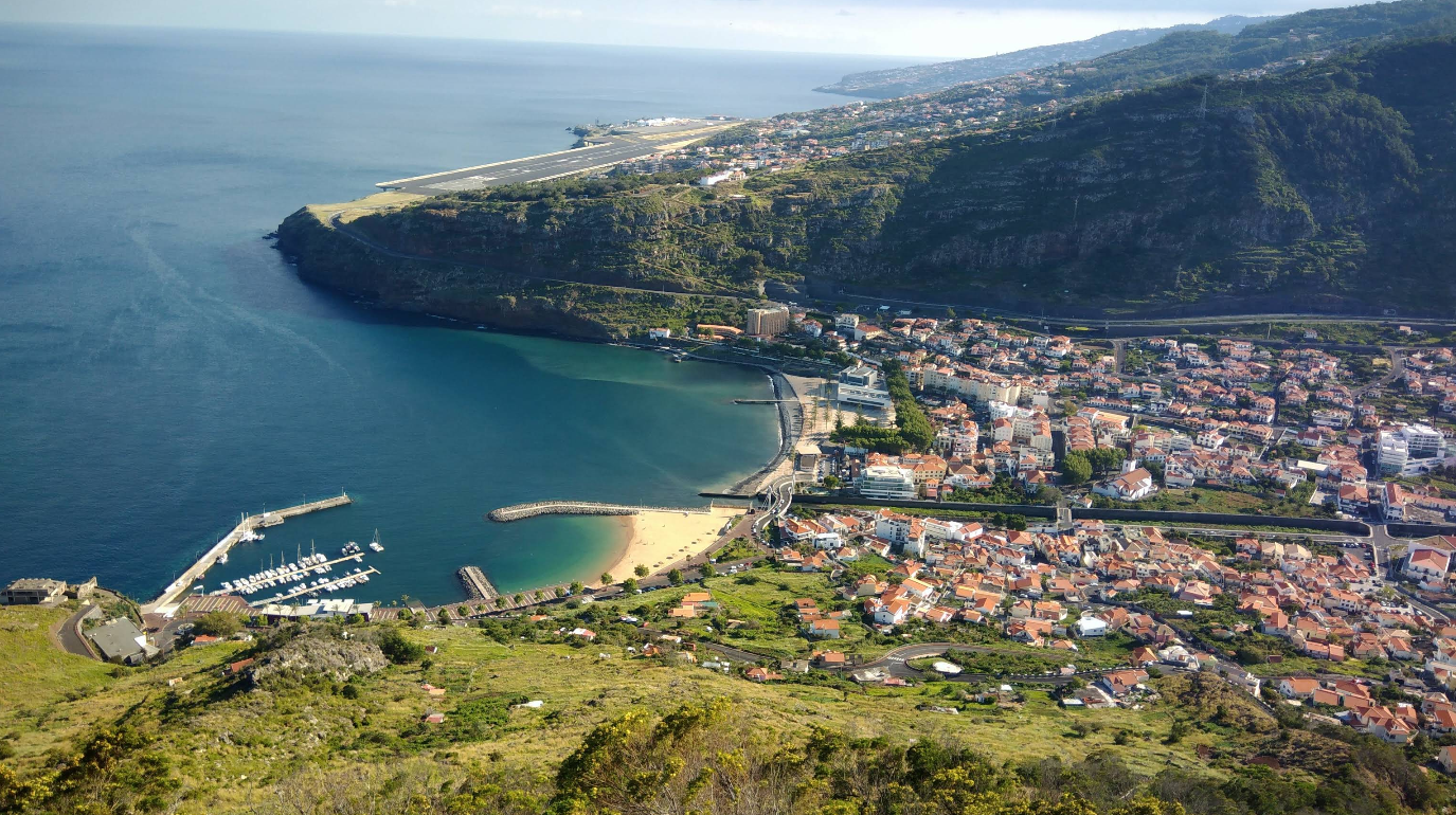 Aerial view of a coastal town with a marina, sandy beach, and a peninsula. Buildings cluster near the shore, with green hills and an airport runway visible in the background.