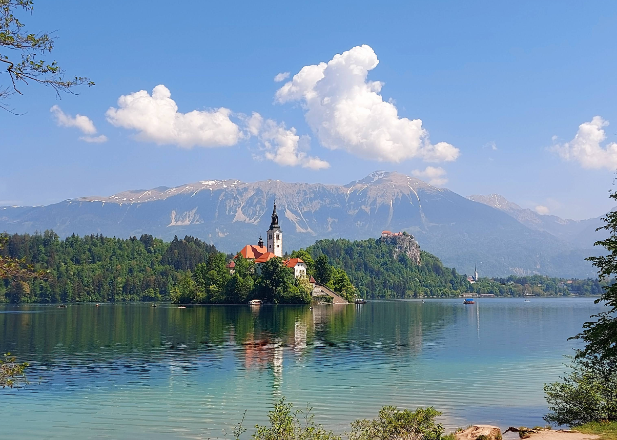 Lake with a small island featuring a church, surrounded by trees and mountains. The water reflects the island and a blue sky with clouds.